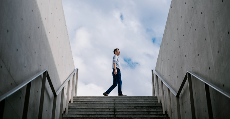 A person walking at the top of an outdoor staircase framed by two concrete walls, under a cloudy sky.